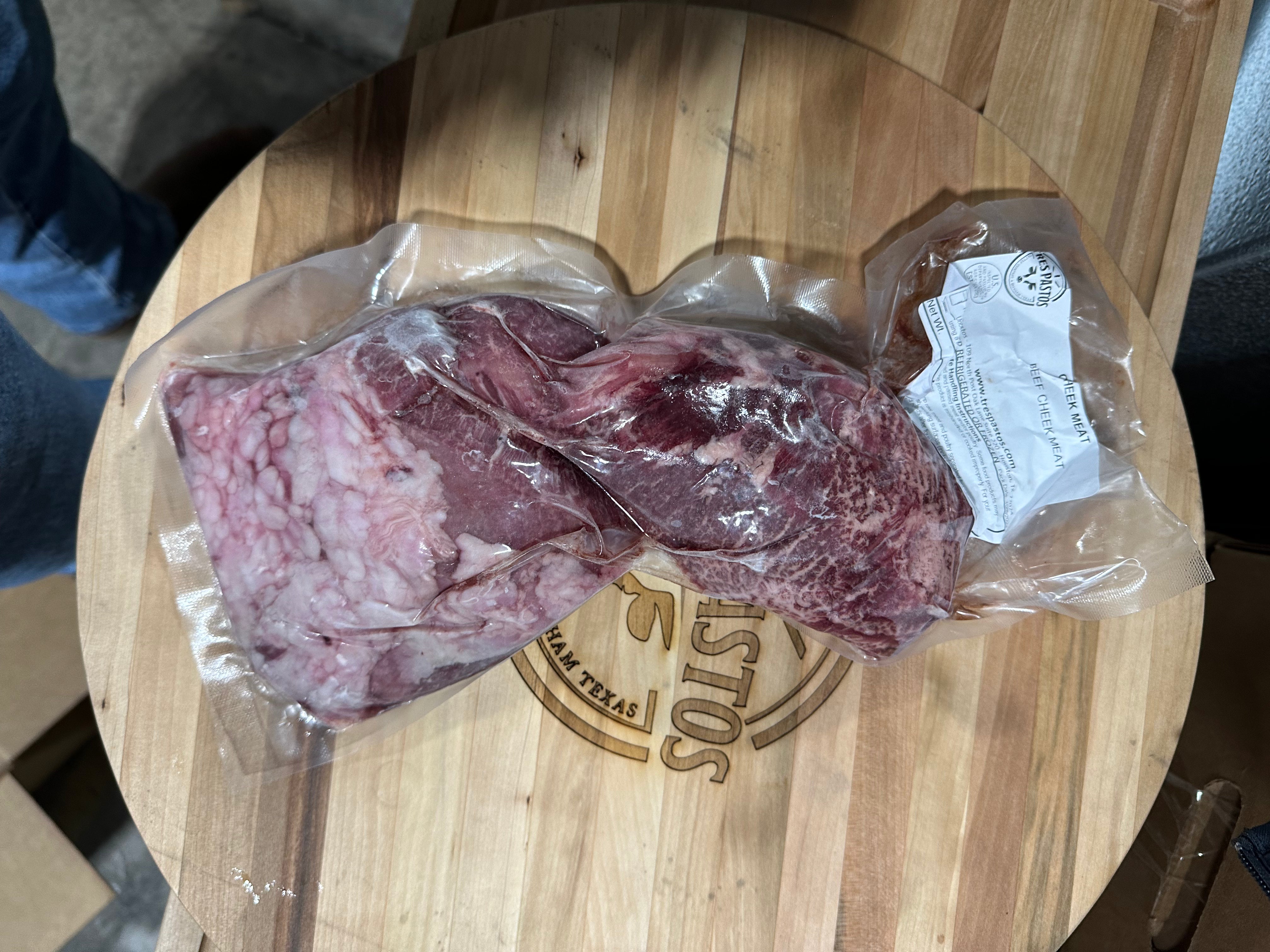 A vacuum-sealed plastic bag containing fresh beef cheek meat, placed on a wooden cutting board.