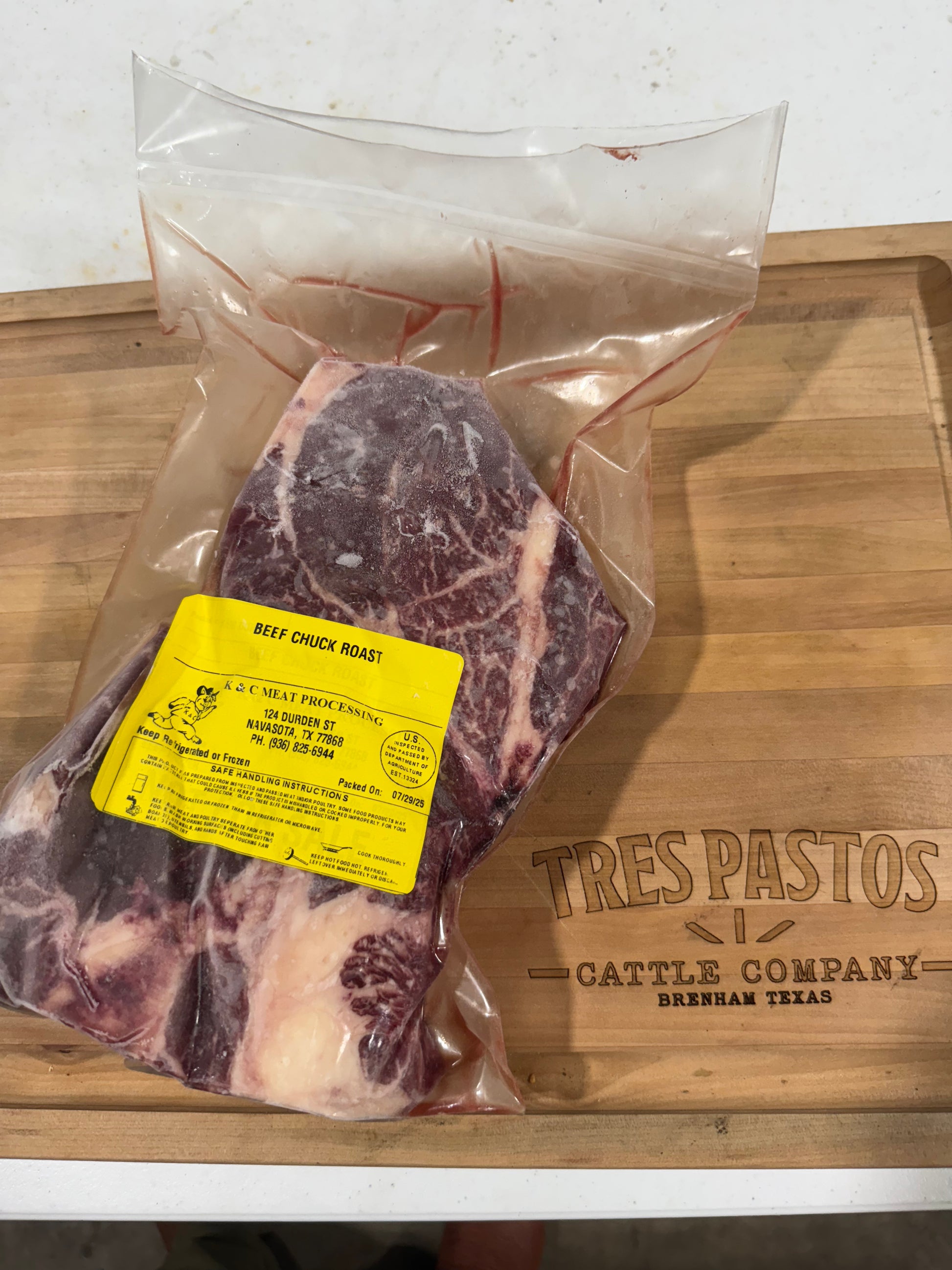 Raw beef chuck roast in a clear plastic bag with a yellow label on a wooden cutting board with 'Tres Pastos Cattle Company' branding.